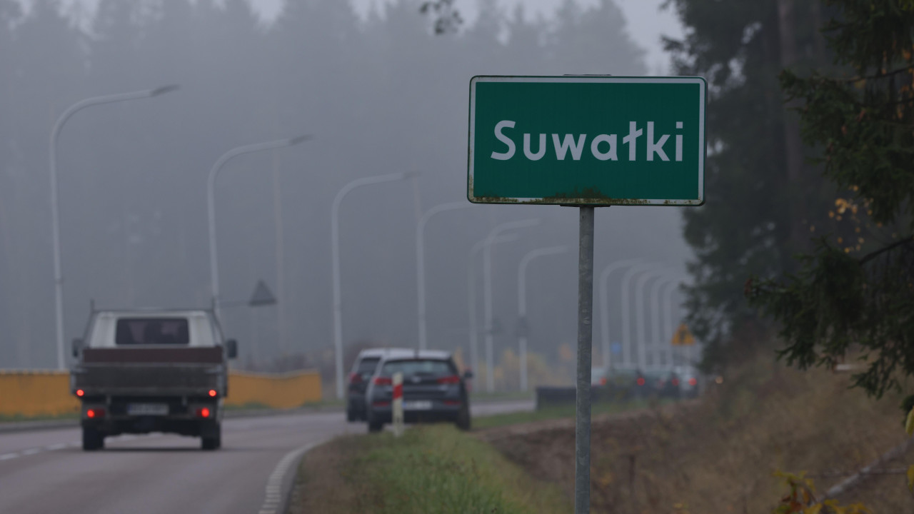 Dva automobila prolaze pored grada na putu sa zelenim saobraćajnim znakom za Suvalki u Poljskoj. U pozadini se nalaze drveće i ulična rasveta