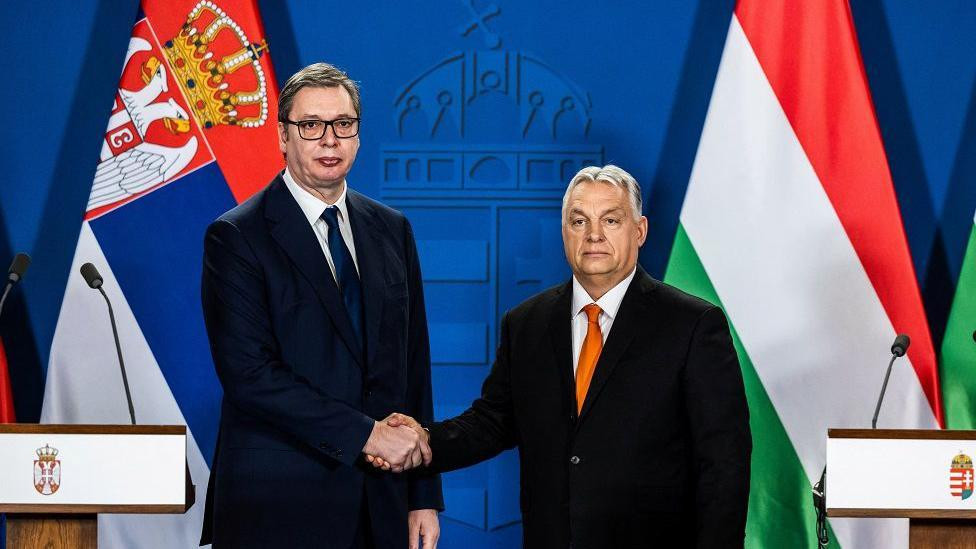 Vučić i Orban u odelima se rukuju smrknutih izraza lica sa zastavama Srbije i Mađarske u pozadini
