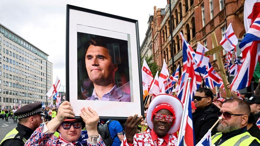 protest u londonu, čarli kirk, charlie kirk, muškarac u odelu u bojama zastava velike britanije nosi sliku sa likom Čarlija Kirka ubijenog konzervativnog aktiviste iz Amerike