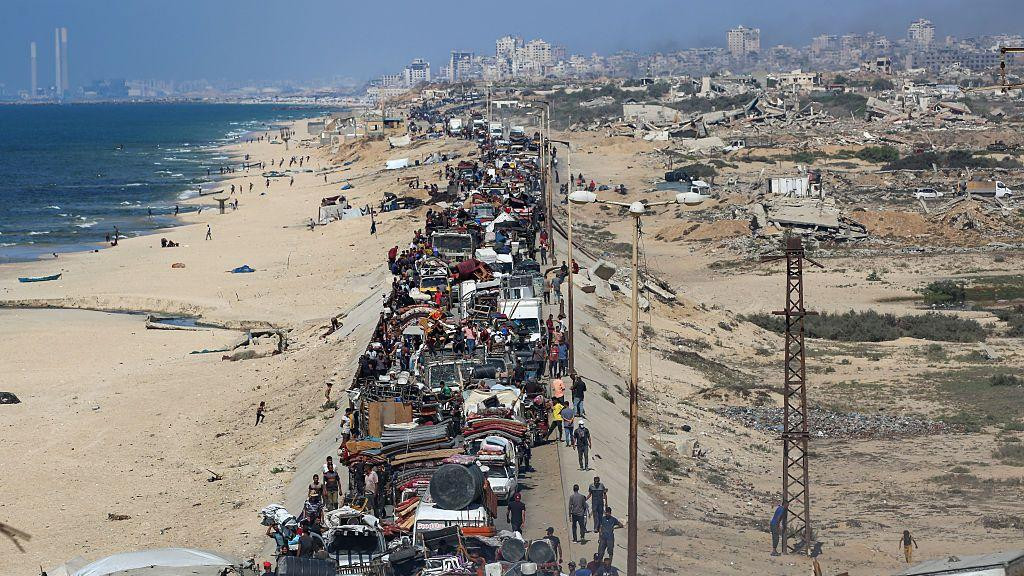 Gomila ljudi putuje priobalnim putem automobilima natovarenim njihovim stvarima. Predeo je neplodan, a uništene zgrade se vide u daljini.