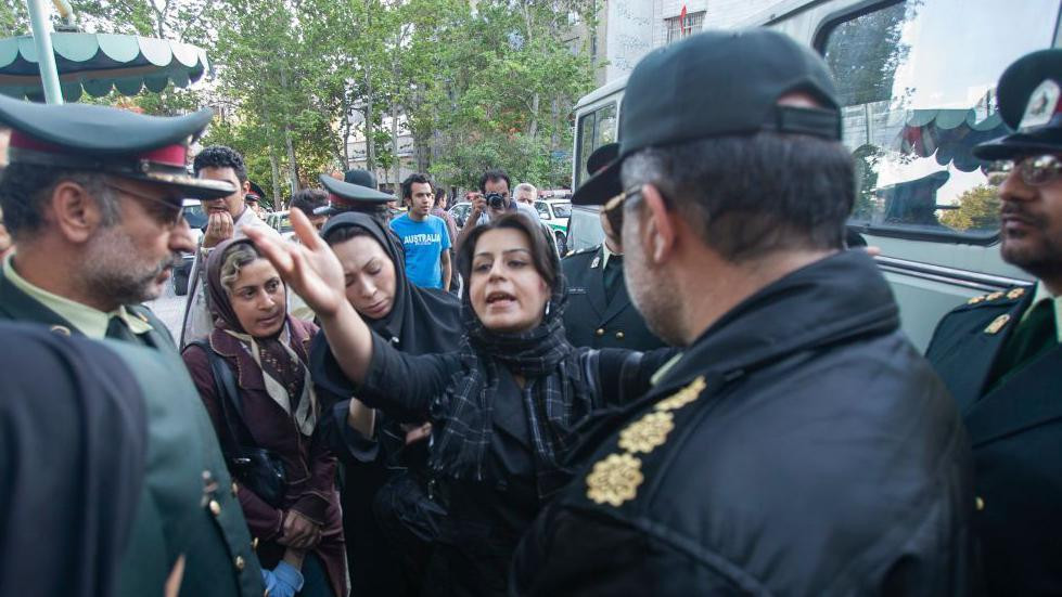 Policija gleda u ženu koja je podigla ruku i objašnjava dok iza nje stoje dve žene i okupljeni muškarci