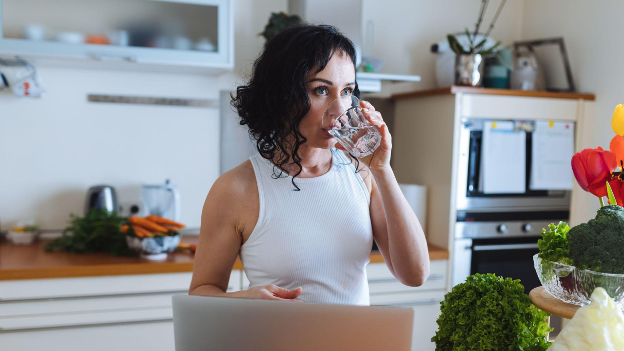 Žena u beloj majici pije vodu u kuhinji, okružena svežim povrćem i laptopom