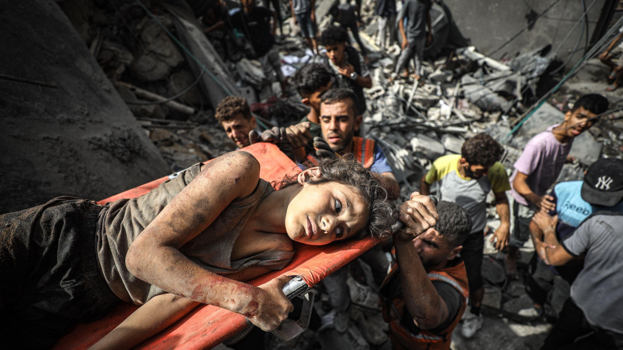 Mlada devojka leži na nosilima dok je izvlače iz uništene zgrade, ali je spasilačka služba izvlači. Drži se za stranu nosila i izgleda očajno.