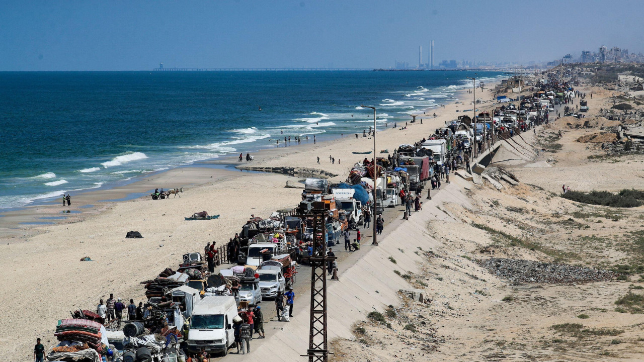 Dugačka kolona automobila u zastoju na priobalnom putu u Gazi, dok ljudi peške obilaze vozila. Obala se vidi levo od slike, a soliteri u daljini.