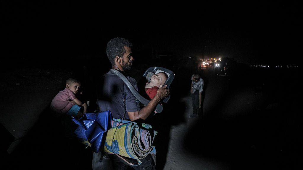 Čovek beži sa decom peške. Nosi dete koje spava u auto-stolici prebačenoj preko ramena, a na leđima nosi tepih. 