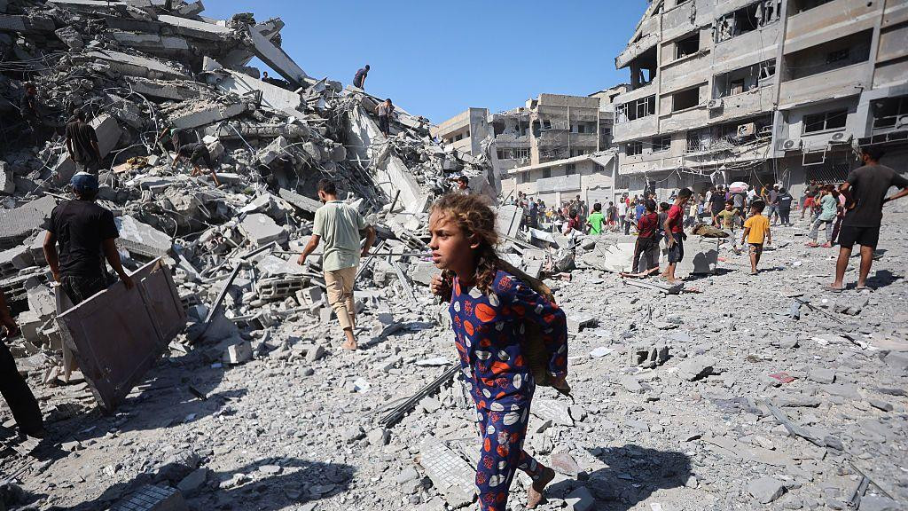 Devojčica u šarenoj plavoj pidžami sa crvenim i belim crtežima hoda bosa po ruševinama dok raseljeni Palestinci spasavaju predmete iz ruševina kule Al-Gafri nakon što je uništena u izraelskim napadima.