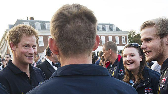 Princ Hari okružen ljudima. Muškarac u jakni na kojoj piše Britanske oružane snage gleda u njega a leđima je okrenut kameri  