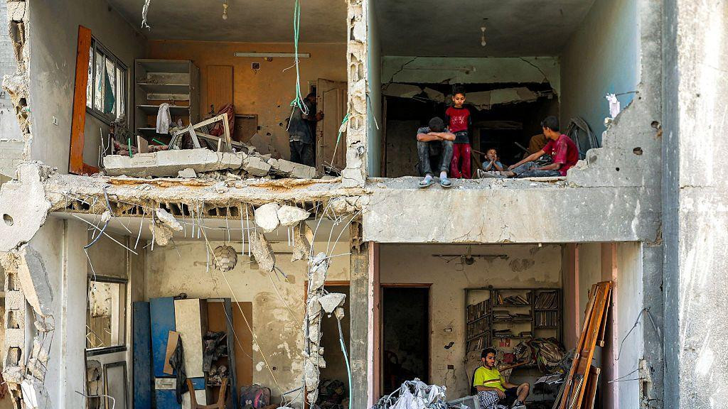 Čovek i deca sede u sobama u teško oštećenoj zgradi. Čitav prednji deo zgrade nedostaje, ostavljajući vidljive četiri sobe, sa ruševinama razbacanim po celoj zgradi.