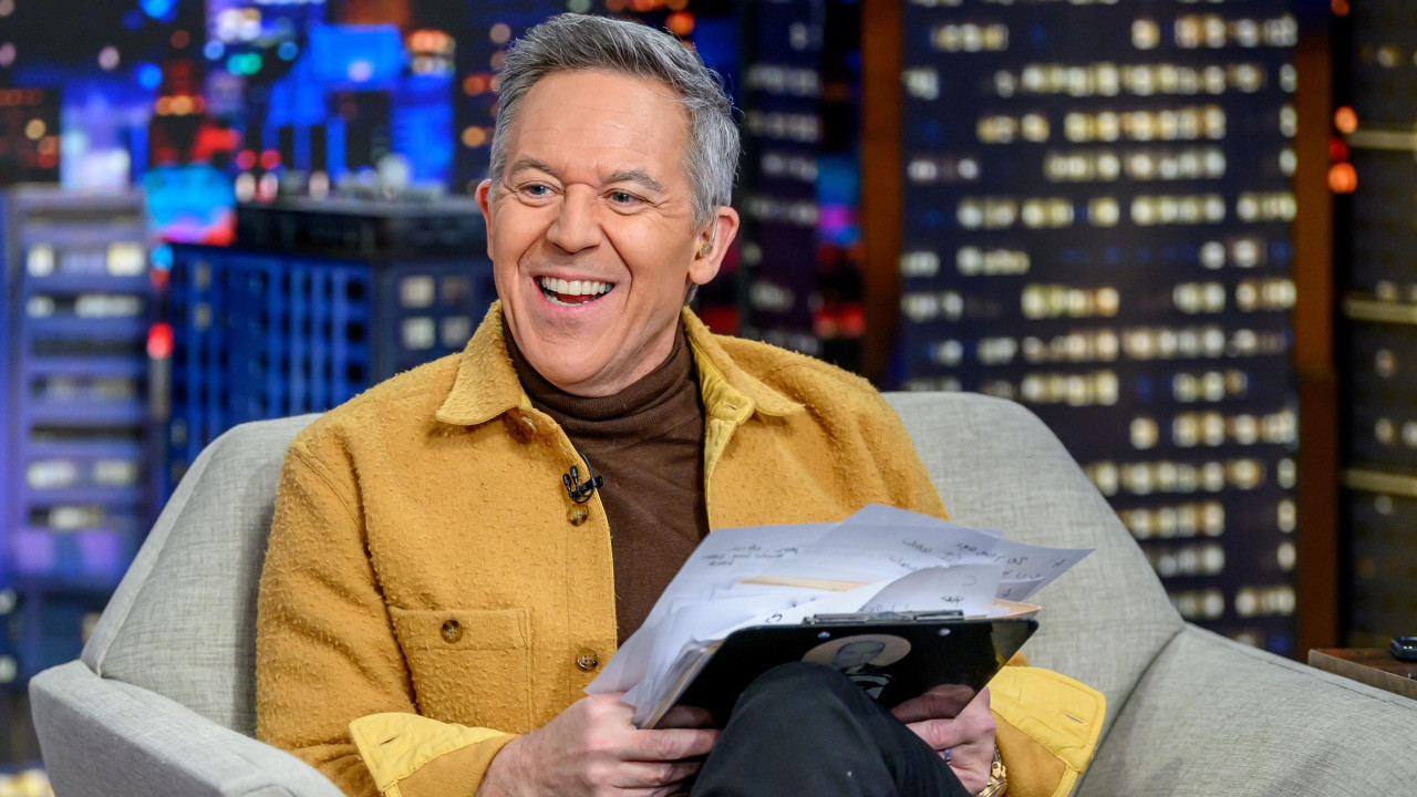 Greg Gutfeld sitting in a chair and smiling against a nighttime cityscape backdrop on the set of his Fox News show