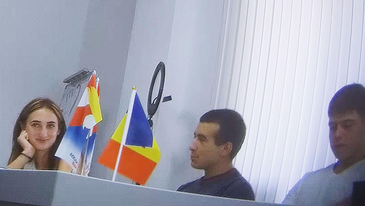 
                    Žena sa dugom braon kosom (Alina Juk) smeši se grupi muškaraca koja je okružuje. Svi sede za dugačkim stolom na kome su dve zastava, od kojih je jedna moldavska sa crveno