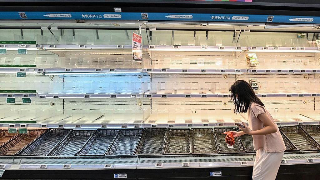 Žena stoji sa kesom crnog luka ispred prazne police u supermarketu