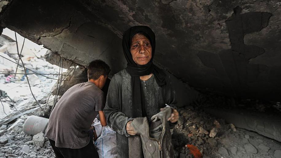 Žena u sivoj odeći sa crnom maramom na glavi stoji ispod ruševine. Iza nje dečak