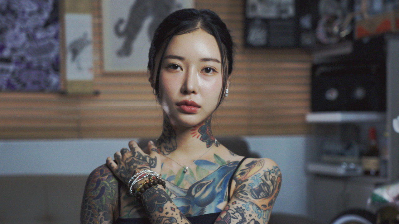 Headshot of Ahn Lina with her hair tied up and her hand on her shoulder, with her arms, shoulders and neck covered in tattoos.