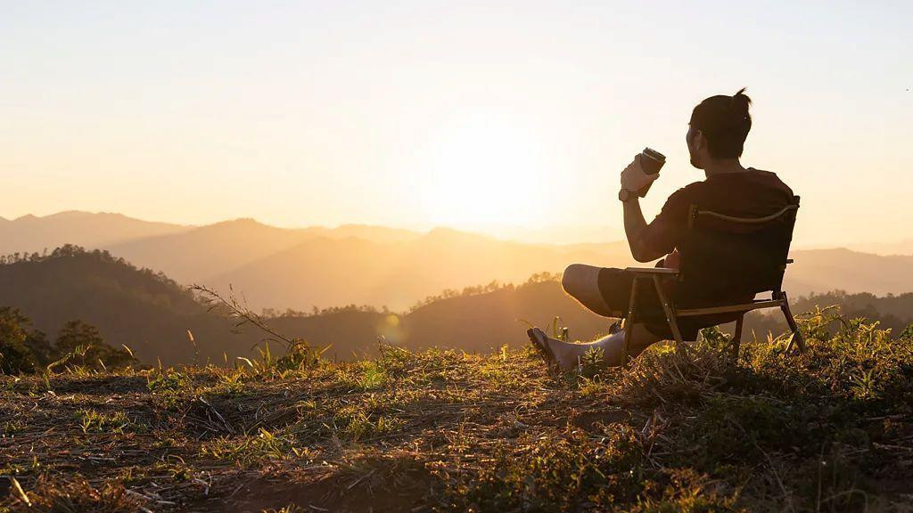 muškarac sedi na stolici na uzvišenju okrenut kameri, u ruci drži kafu dok gleda u sunce koje izlazi
