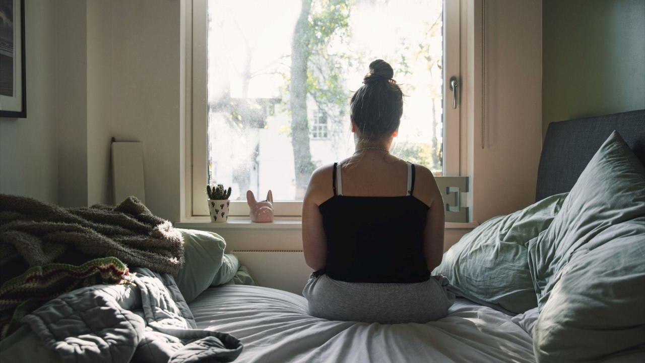 Tinejdžerka gleda kroz prozor dok sedi na krevetu u majici na bretele