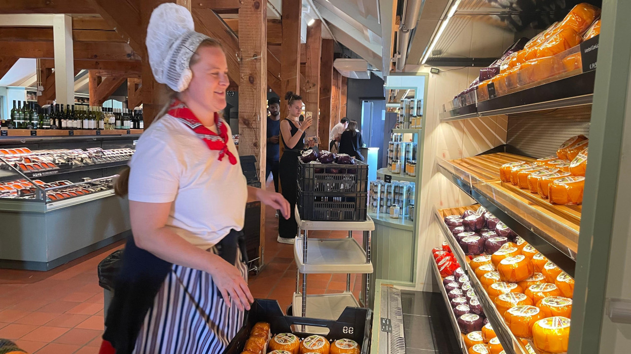 žena obučena u tradicionalnu nošnju - bela majica i bela kapica, sa crvenom maramom oko vrata ispred rafova na kojima su poređani sirevi 