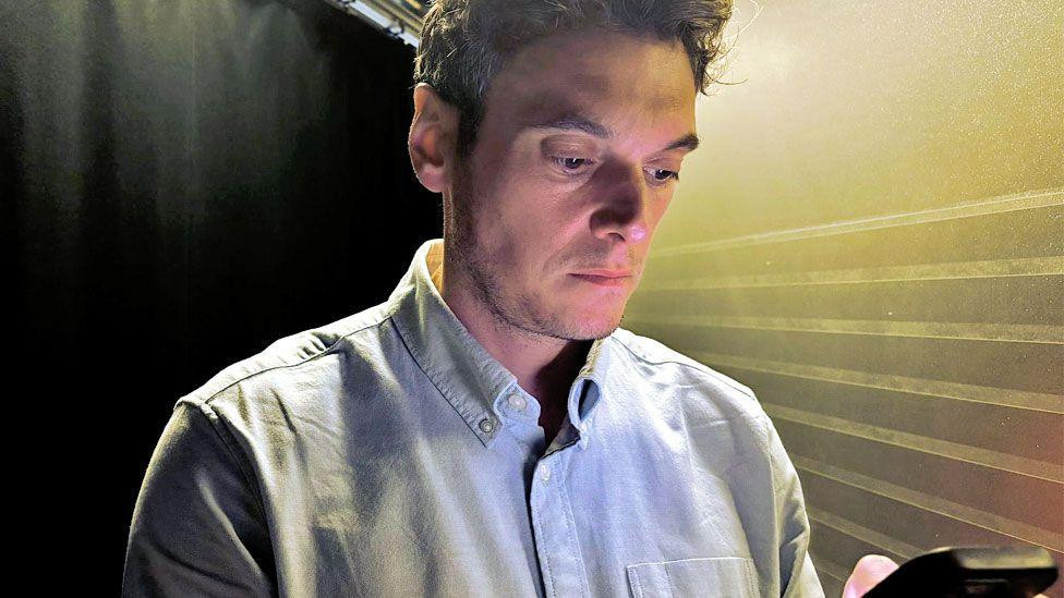 Joe Tidy looking down at a phone. He has short brown hair and is wearing a light blue shirt.