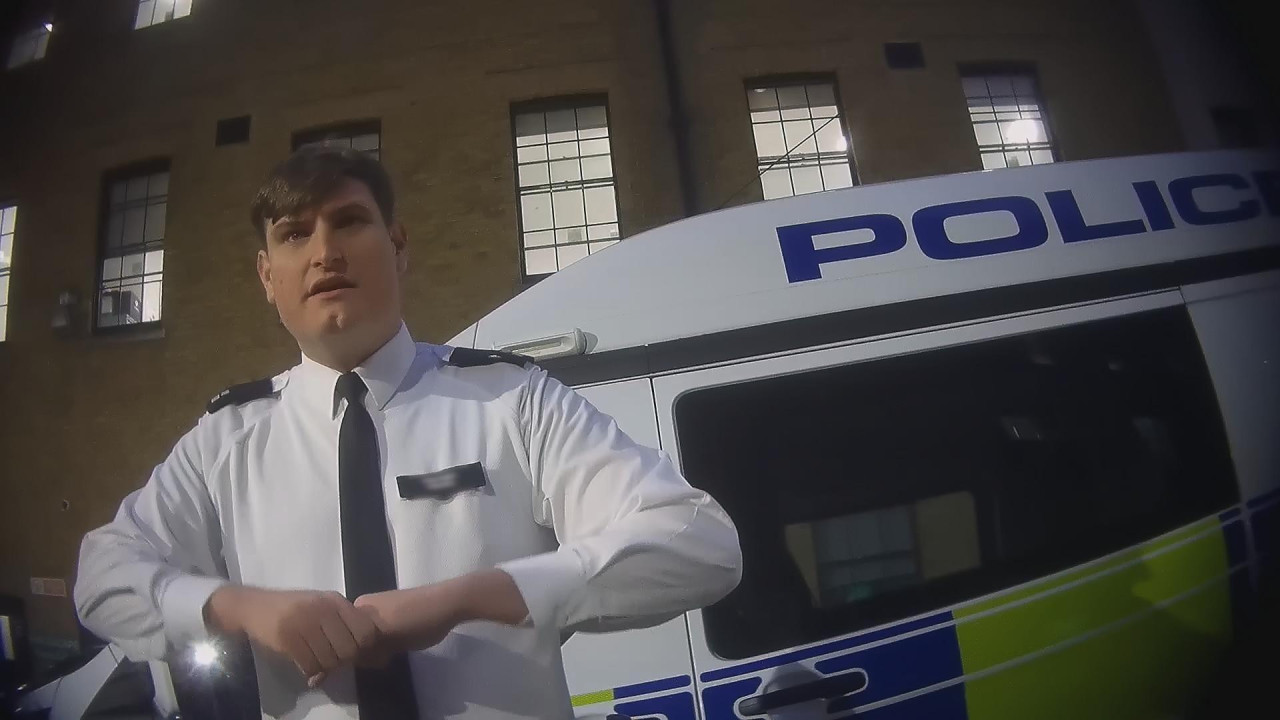 A hidden camera shot, taken slightly from below and at an angle, of an officer in police uniform, a white man with brown hair in a side-parting and a with a cleft chin, standing in front of a police van in the courtyard of Charing Cross police station, with windows of the station all around. He is pulling with his right hand on the fingers of his left as he discusses taking fingerprints by force