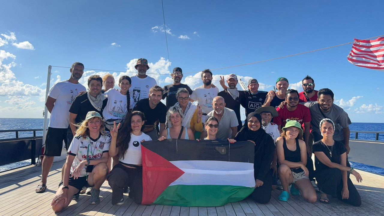 Grupa od oko 25 ljudi pravi selfi na palubi broda po sunčanom danu. Obučeni su ležerno i deluju srećno, osmehujući se kameri. Drže palestinsku zastavu, a nekoliko njih podiže ruke u znaku „v kao pobeda“. Crveno-bela zastava se vidi kako se vijori na vetru desno od slike.