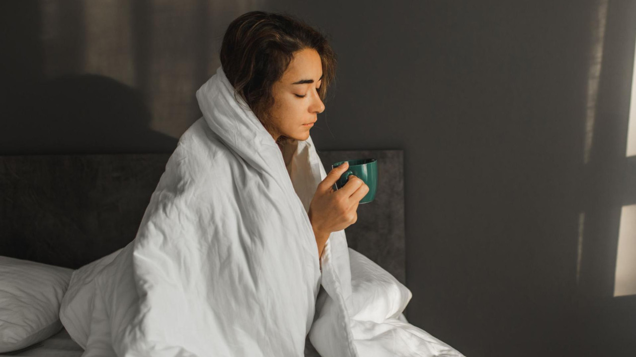 grip, žena ima grip, devojka ima grup, žena sedi pored kreveta umotana u beli jorgan, dok drži zelenu šolju. Ima tamnu kosu i pogled joj je spušten ka podu