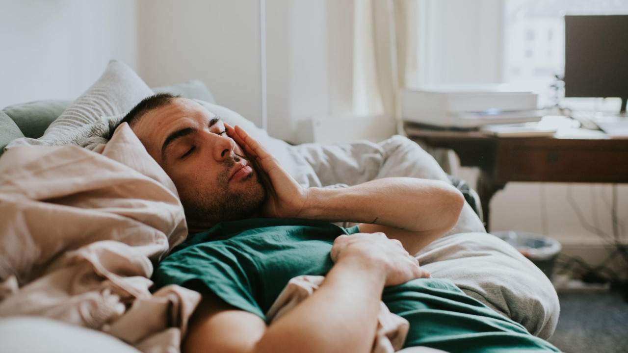 Čovek leži u krevetu držeći levu stranu lica rukom. Nosi tamnozelenu majicu
