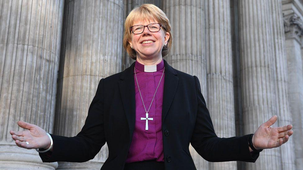 
                    Dame Sarah Mullally is dressed in a dark jacket and is wearing a white clerical collar on a purple shirt. Her arms are outstretched and she is smiling. Large Roman style 