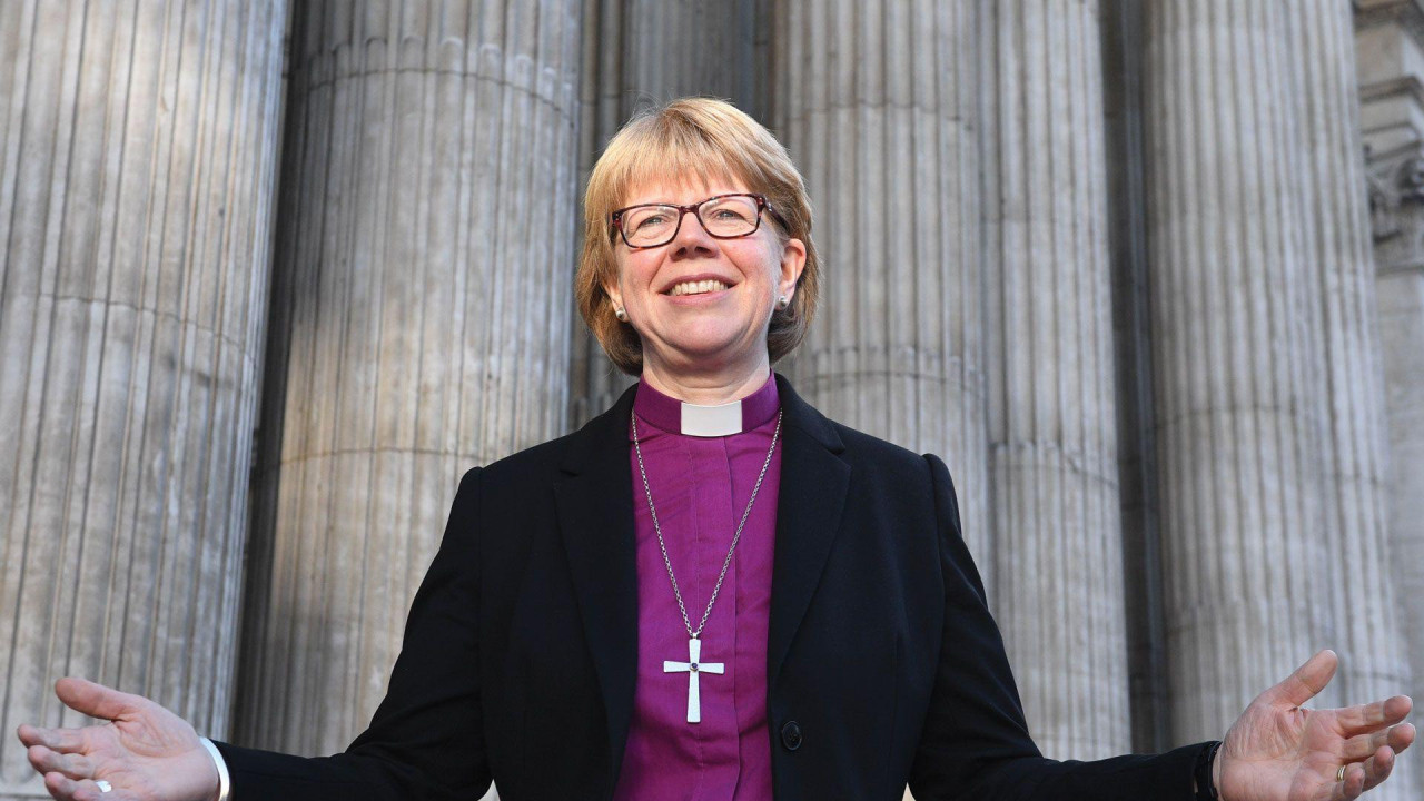 Sara Malali u crnoj odori i ljubičastoj košulji, sa okačenim krstom preko, širi ruke i smeke se, ima plavu kosu i crne naočale