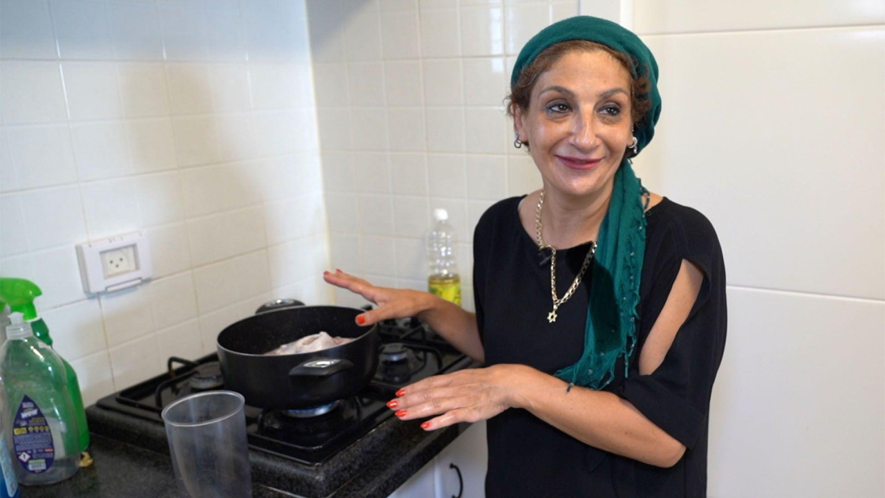 Žena stoji pored otvorenog crnog lonca na šporetu. Nosi zelenu maramu oko glave, crnu majicu i zlatnu ogrlicu sa zvezdom. Iza nje je boca ulja, a zidovi su prekriveni belim pločicama