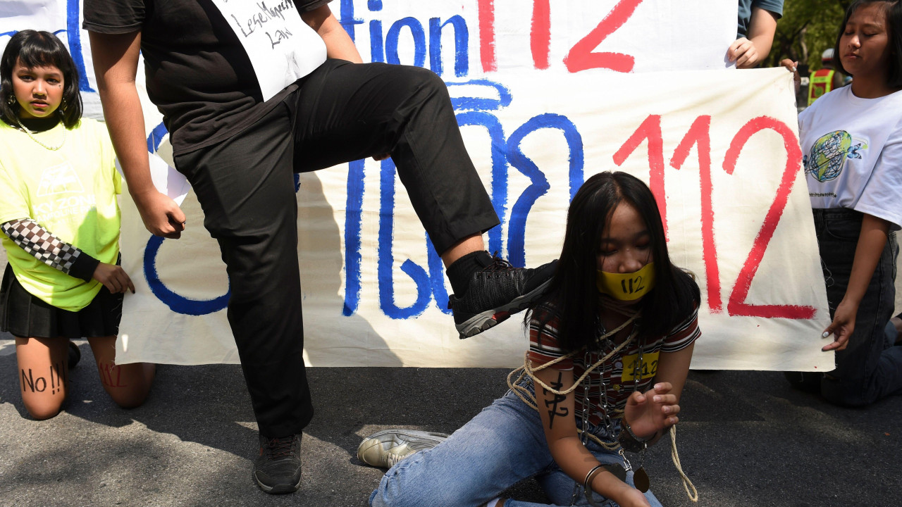 Demonstrant stavlja nogu na rame mlade demonstrantkinje koja kleči na zemlji vezana konopcem. Na transpatrentu iza nje je broj 112, član tajlandskog zakona koji kriminalizuje vređanje monarha