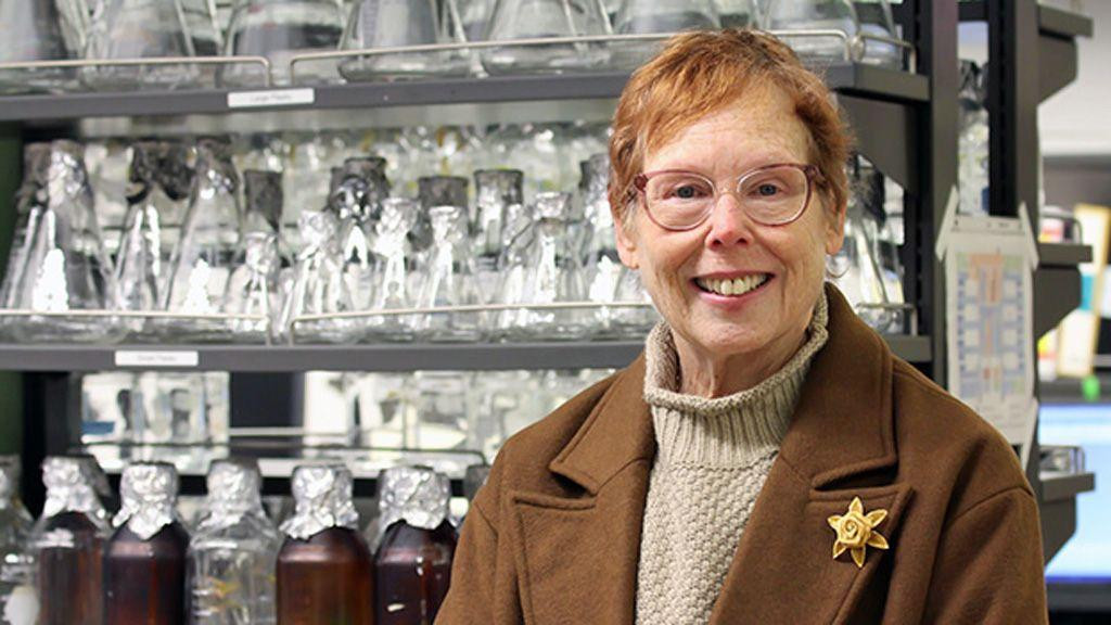 Fotografija doktorke Nore Disis u laboratoriji. Ima kratku crvenu kosu i nosi naočare sa crvenim okvirom, džemper bež boje i kaput boje braon boje. Iza nje su metalne police sa staklenim bocama.