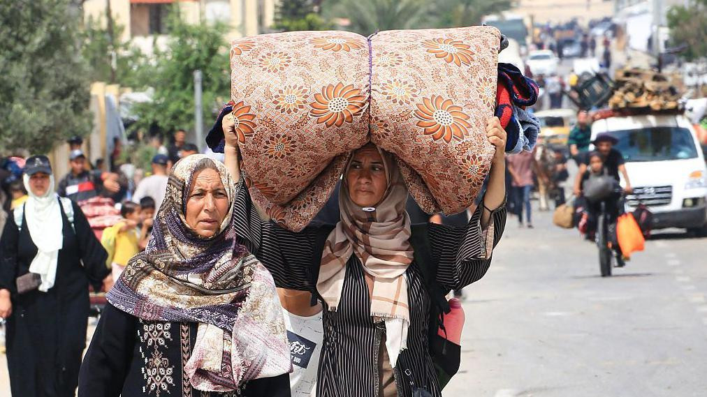 Dve žene sa maramama na glavi, jedna od njih nosi smotani dušek na glavi, u daljini natoverana kola stvarima