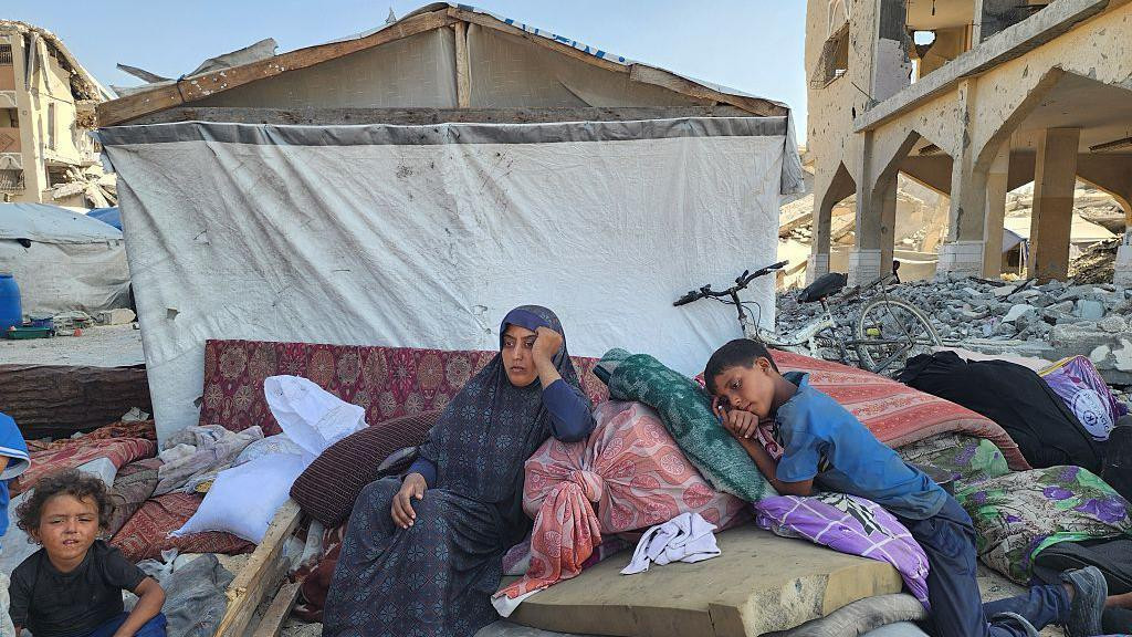 Žena sedi između dvoje dece, jednog predškolskog uzrasta i starijeg dečaka. Oni se smeštaju na gomilu dušeka i stvari ispred improvizovanog skloništa napravljenog od platna i komadića trave. Stariji dečak se oslanja na jastuk kao da pokušava da zaspi, žena naslanja glavu na ruku sa tužnim, umornim izrazom lica. Ostaci uništenih zgrada mogu se videti sa obe strane skloništa. Snimljeno u oblasti al-Mavasi, 17. septembra 2025.