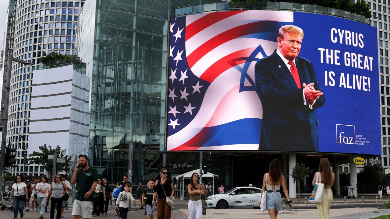 Ljudi šetaju ulicama Tel Aviva dok iznad njih na ekranu slika Donalda Trampa iza kojeg su američka i izraelska zastava