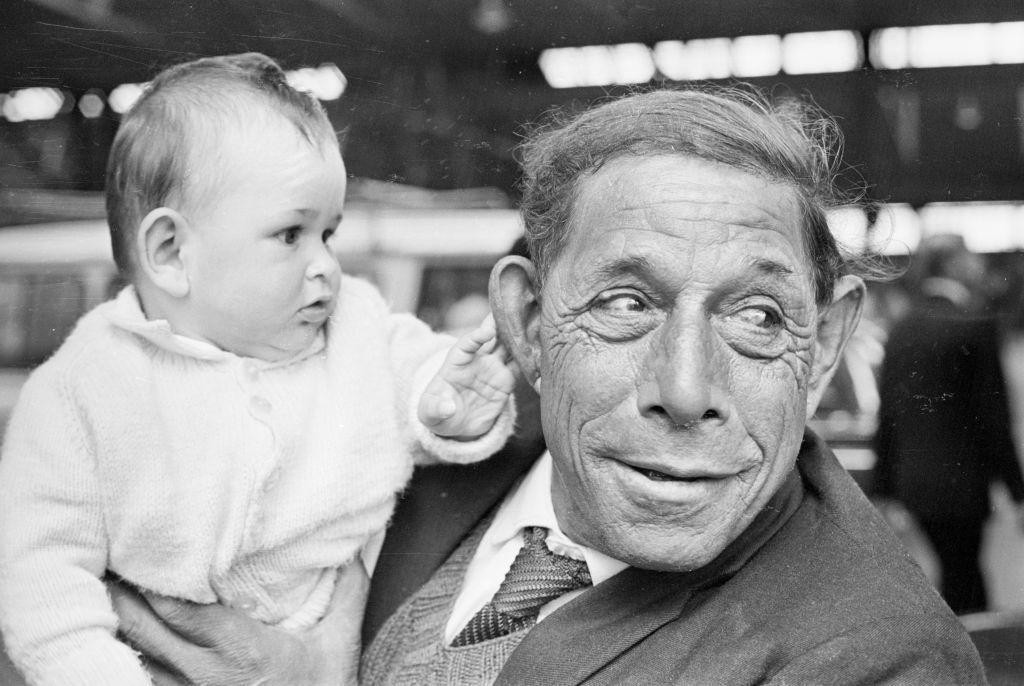 
                    A baby in a white cardigan gently touches the ear of the man holding him. The man, dressed in a jacket and tie, looks away from the child. Both are among the Tristan da C