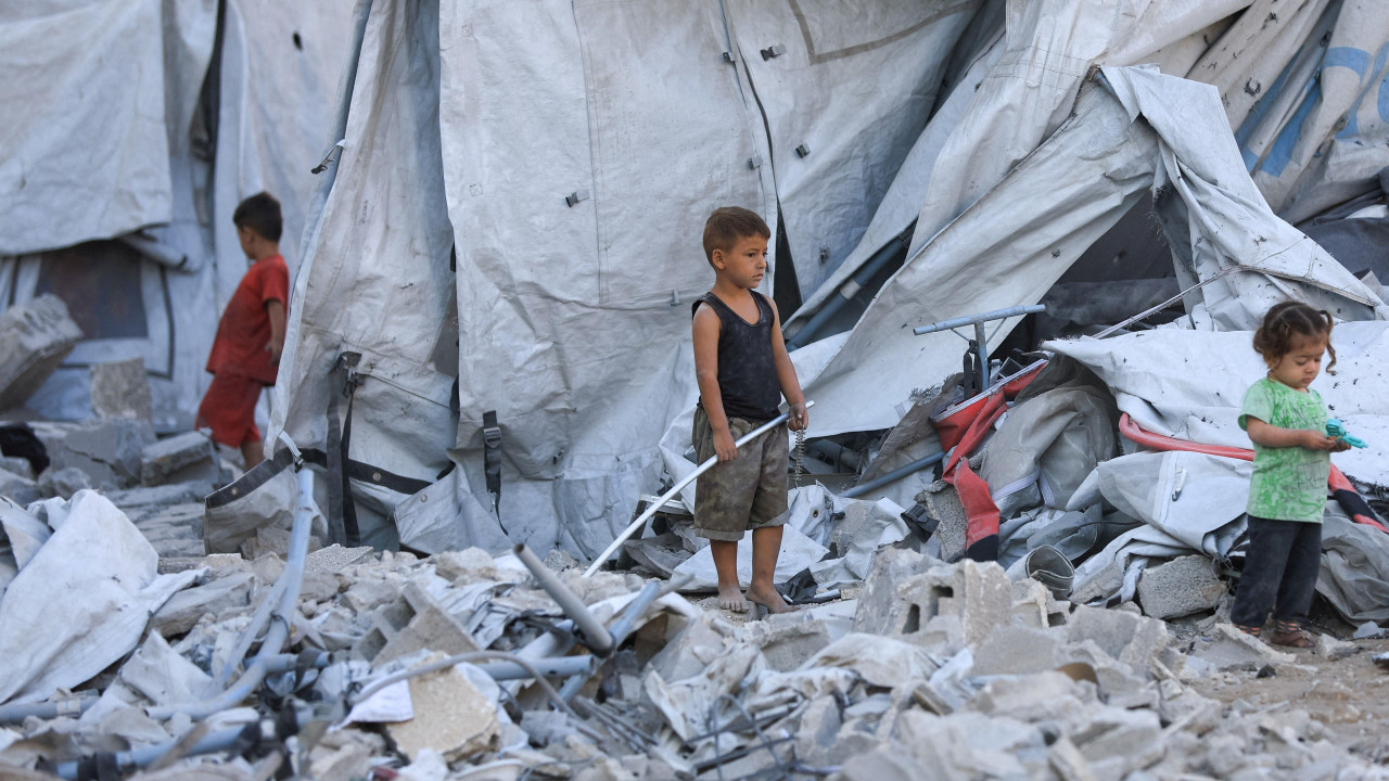 dva dečaka i devojčica stoje u ruševinama među šatorima u gradu Gazi 