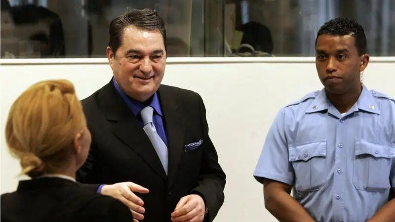 
                    Nebojša Pavković u haškoj sudnici u crnom odelu i plavoj košulji, smeje se i pruža ruku. Pored njega je policajac u uniformi, stoji sklopljenih ruku. Ispred Pavkovića je 