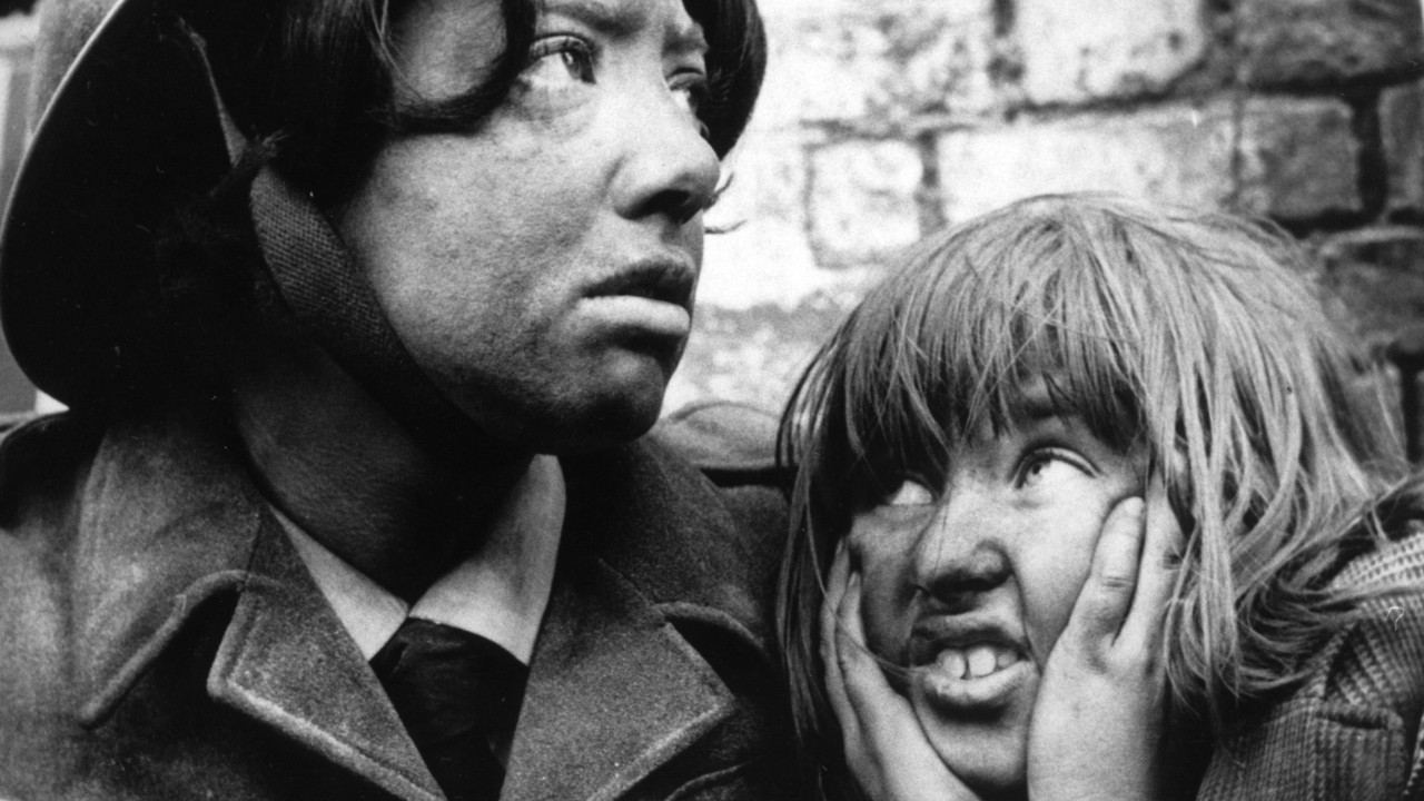 Crno bela fotografija na kojoj su mlada žena u uniformi i devojčica koja rukama drži uši zatvorene. Lica obe deluju prljavo. 