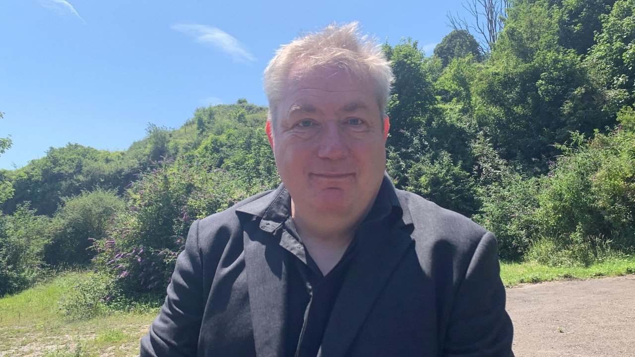 Sredovečni čovek sa sedom kosom, obučen u crnu košulju i sako, osmehuje se. Stoji napolju ispred nekog žbunja po sunčanom danu.