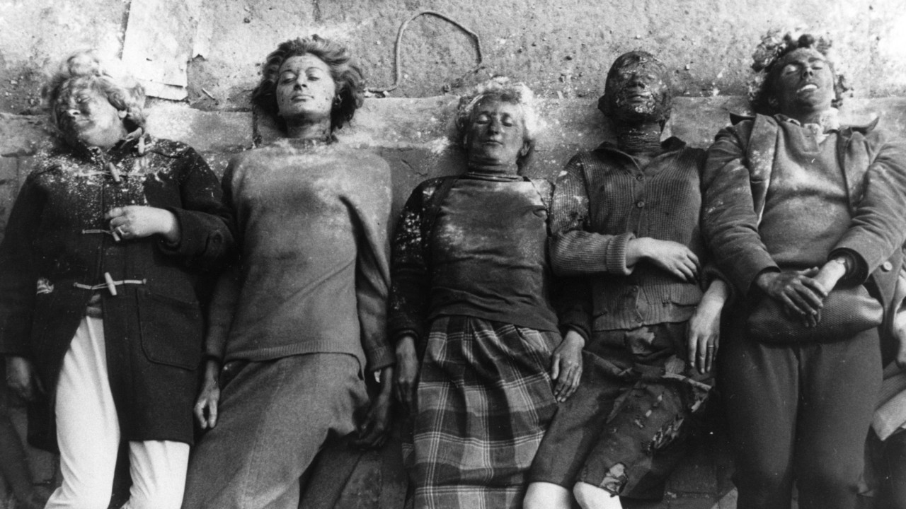 A black and white image of five people in clothing from the 1960s lying on the ground, appearing dead. Their faces have been made to look disfigured by make up. 