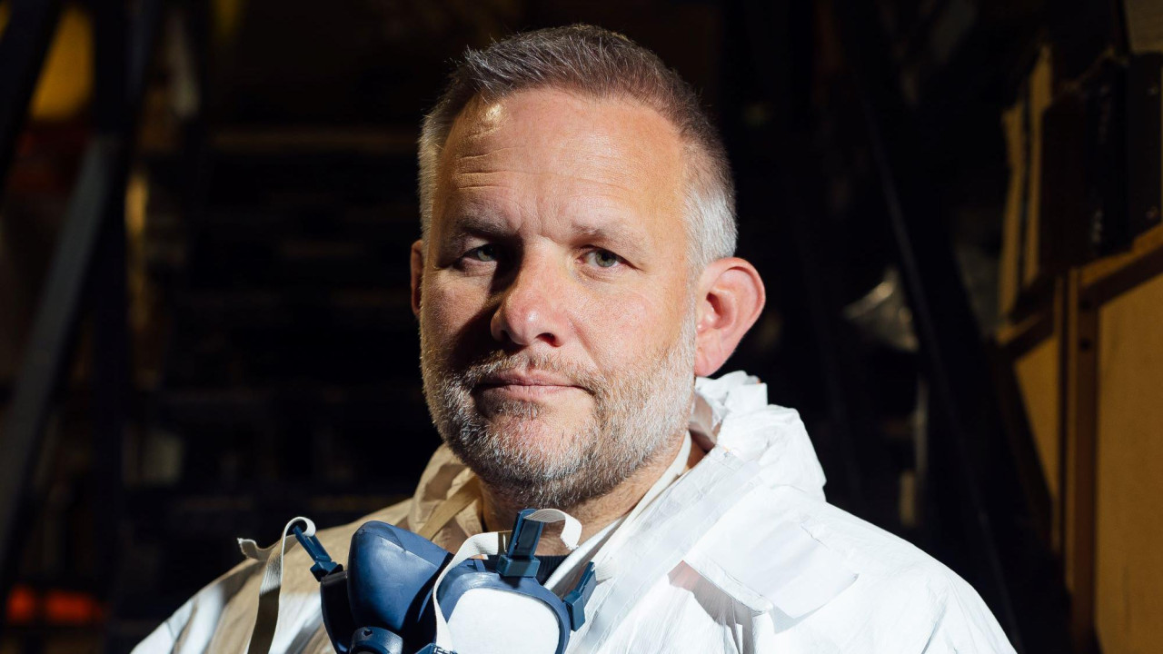 Fotografija Bena Džajlsa izbliza. Ima 49 godina, kratku sedu kosu. Nosi masku za lice oko vrata. Nosi zaštitnu opremu oko vrata, belo odelo sa kapuljačom