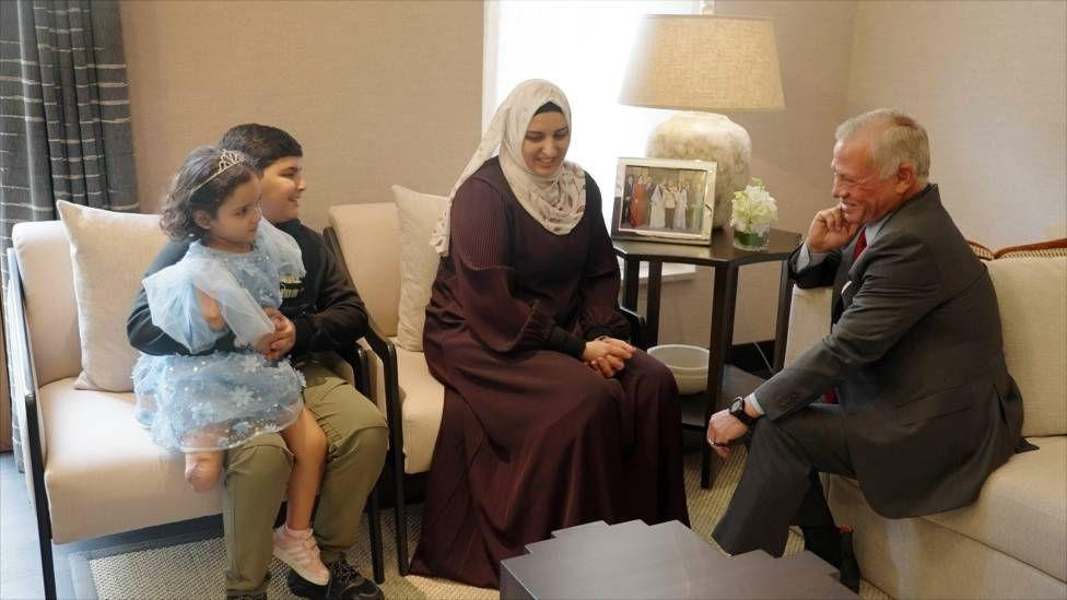 A young girl, missing two arms and a leg, sits on her brother's lap, wearing a blue sparkly dress. To the right, her mother sits smiling, next to a smiling King Abdullah of Jordan.