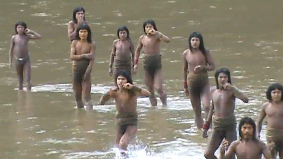 
                    A group of Mashco Piro tribesman wading shin-deep in a river, calling out to someone on the other side. They are wearing only loin cloths and appear to be trying to catch
