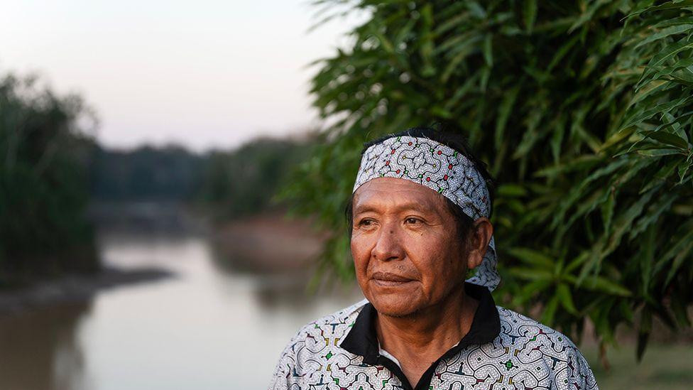 Muškarac nosi šarenu košulju i maramu na glavi, stoji blizu obale reke. U pozadini se vidi mirna voda okružena zelenilom, sa gustim rastinjem sa desne strane i drvećem u daljini