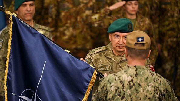 Ulutaş'ın yüzü görünüyor, önde arkası dönük kamuflaj içinde bir asker daha var. En solda nato bayrağı görünüyor. 