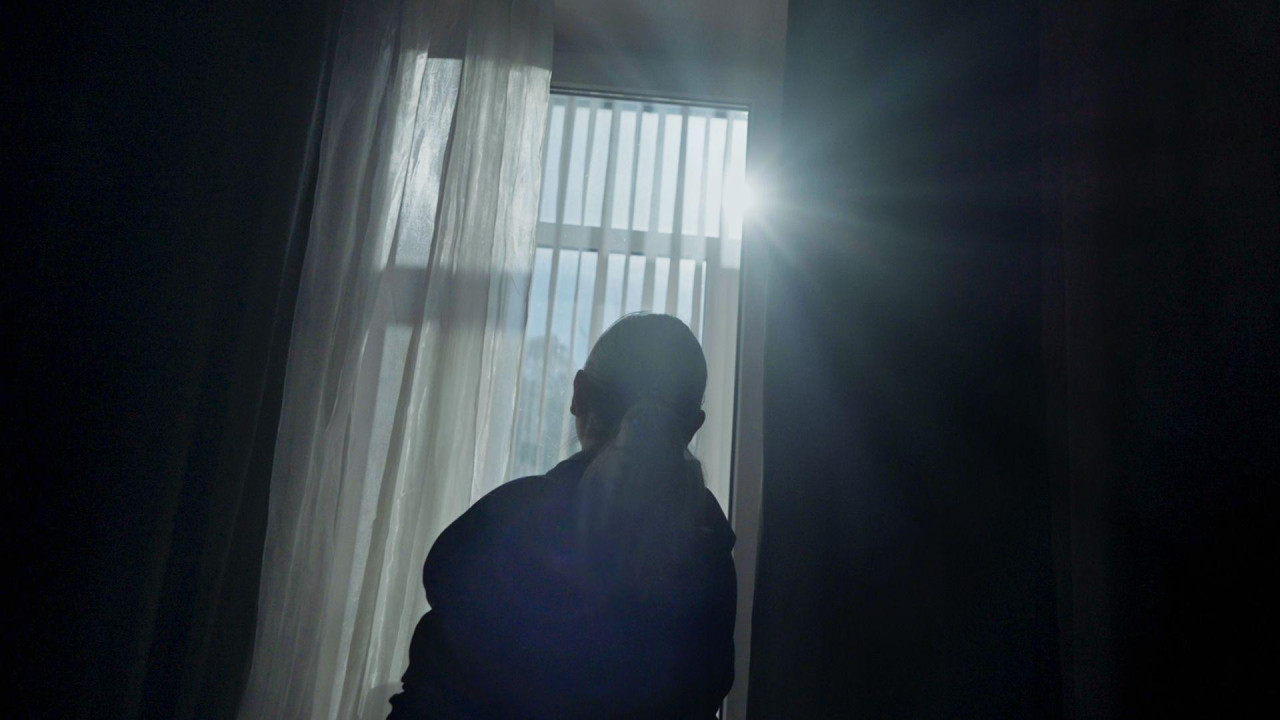 Женщина у зашторенного окна, через которое проглядывает солнце