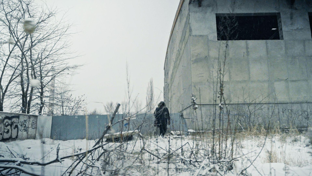 Мужчина с сумкой идет по промзоне зимой мимо здания с темным незастекленным окном в серой бетонной стене