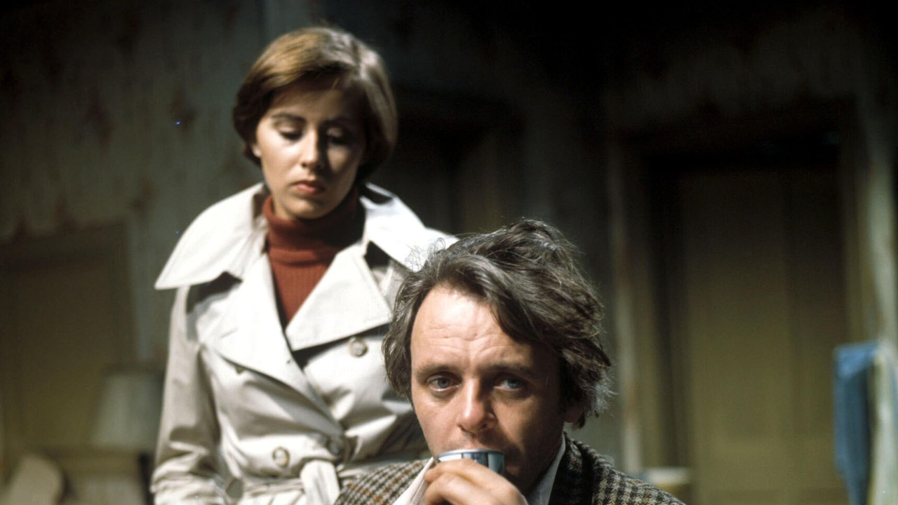 Hopkins sedi za stolom u kariranom sakou, u ruci drži limenku pića, dok je iza njega u pozadini Kejt Neligan u belom dugom mantilu