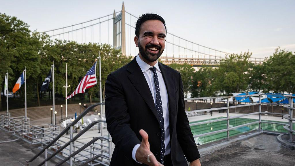 Zohran Mamdani pruža ruku u odelu i beloj košulji