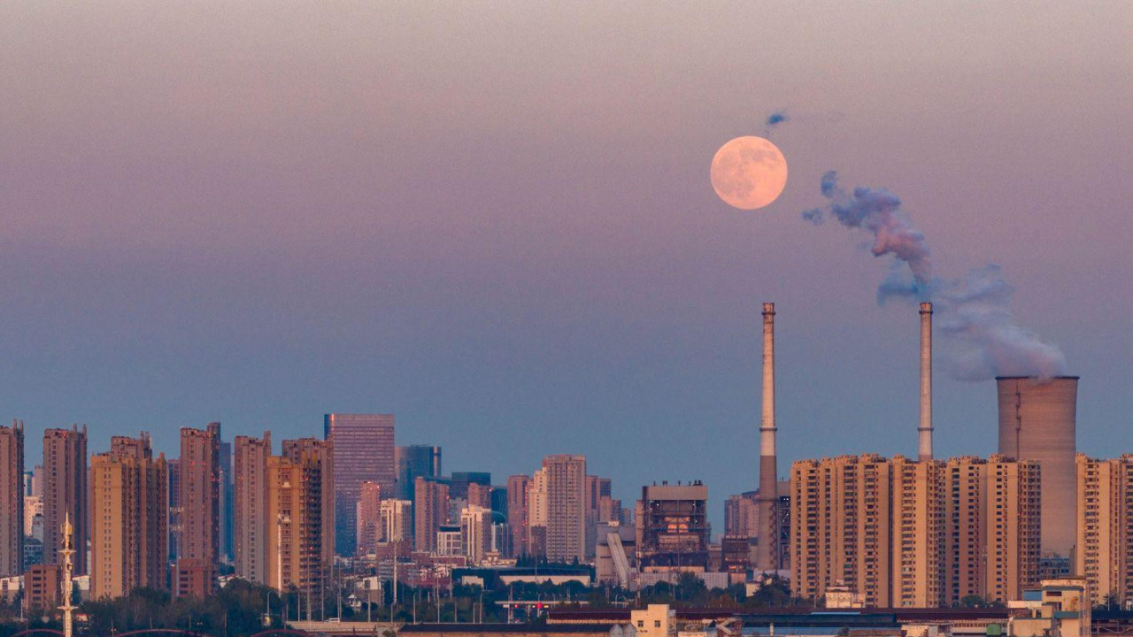 Teretni brodovi se vide kako se kreću kanalom ispred mosta i gradske žičare, dok dimnjaci elektrane dime pored Meseca koji se izdiže iznad grada 