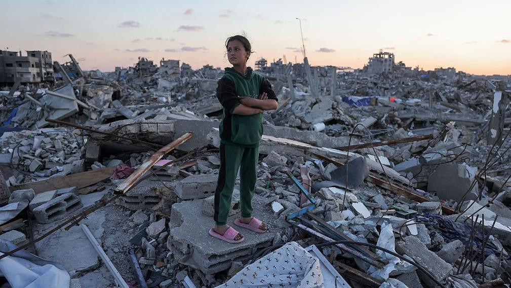 devojčica u zeleno-crnoj trenerci i roze papučama stoji na betonskim ostacima porušenih zgrada na severu Gaze 14. aprila 2025. 